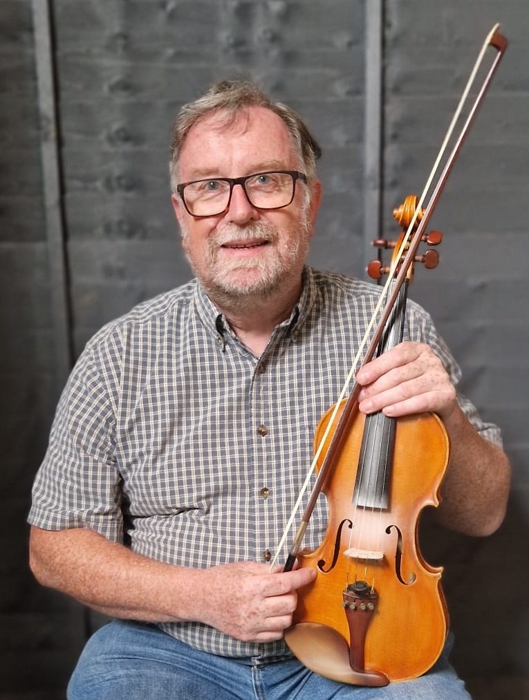 A man wearing glasses, a checked shirt and jeans holding a fiddle.